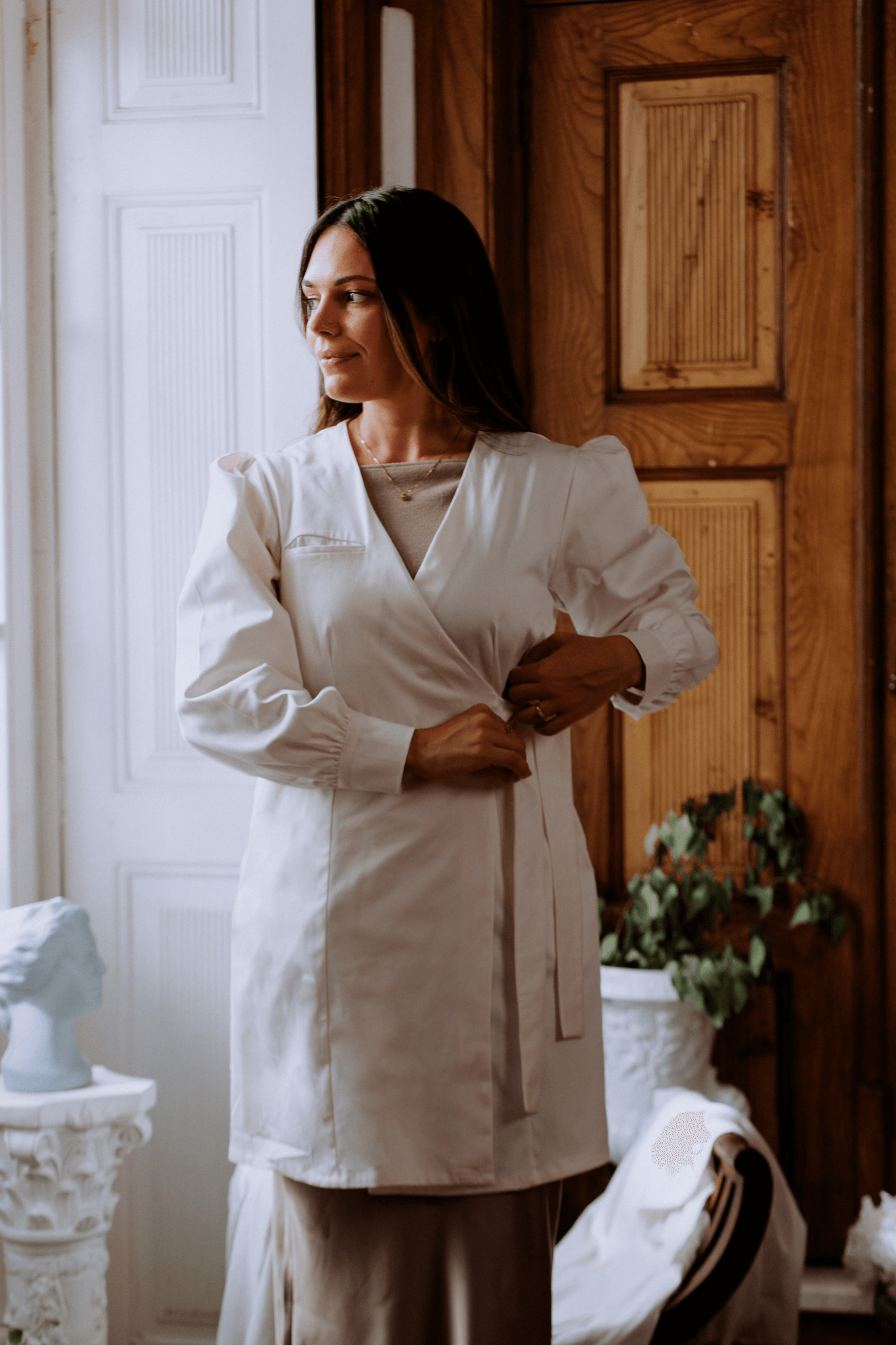 Mulher ajustando bata Agnodice elegante, com mangas volumosas e fecho cruzado, em ambiente clássico.