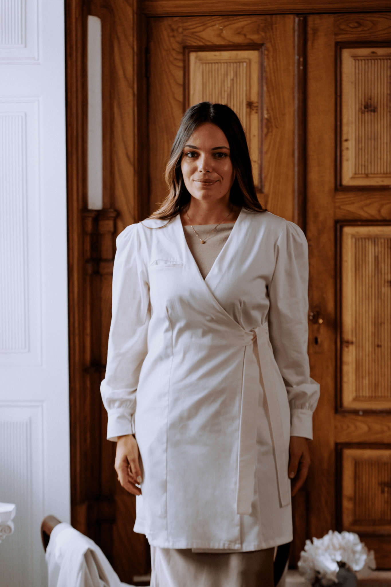 Mulher vestindo bata Agnodice com mangas volumosas e fecho cruzado, em ambiente elegante com madeira ao fundo.