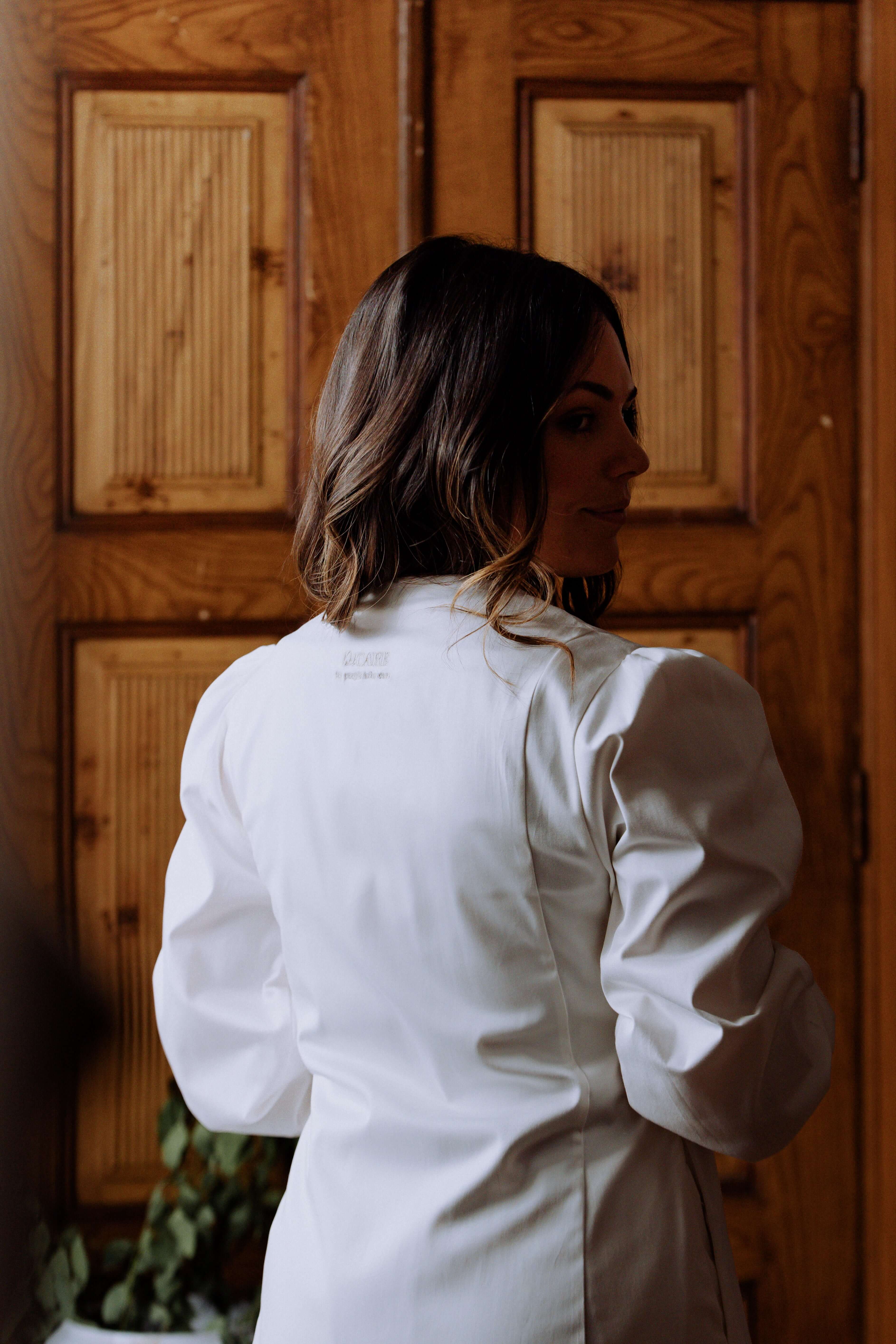 Woman wearing a stylish white bata Agnodice, showcasing voluminous sleeves and a cross closure against a wooden backdrop.
